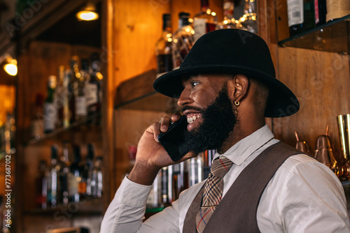 Stylish black bartender in hat using smartphone