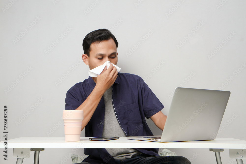 asian man has a runny nose, wipes his nose with tissue paper at workplace while sitting in front of laptop computer. isolated background asian man has a runny nose, wipes his nose with tissue paper at workplace while sitting in front of laptop computer. isolated background