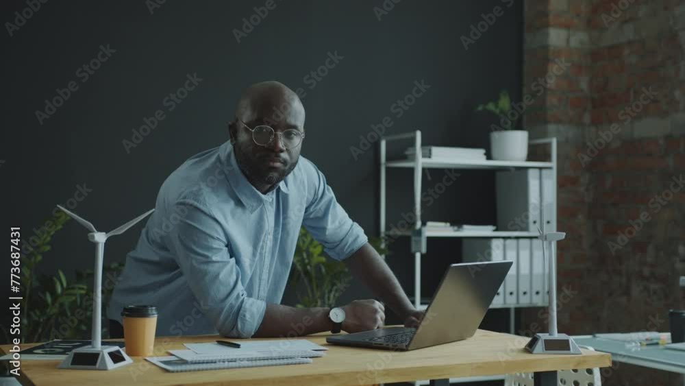 African American energy engineer leaning on desk, working on laptop and posing for camera in the office. Video portrait, medium long shot African American energy engineer leaning on desk, working on laptop and posing for camera in the office. Video portrait, medium long shot