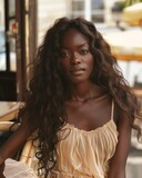 Elegant woman in a beige dress sitting. An elegant African woman with long wavy hair sits in a relaxed pose at a cafe in a chic beige dress