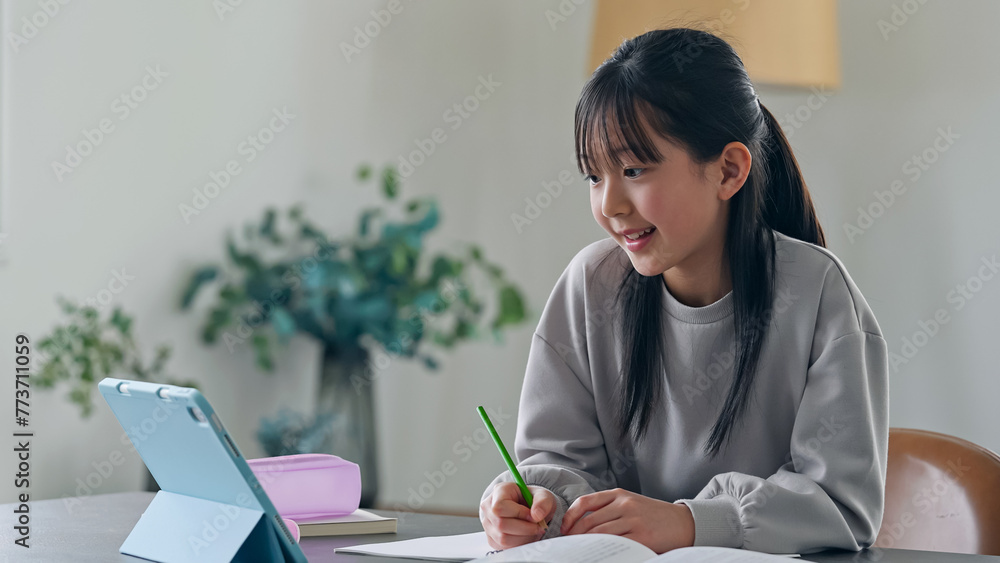 Fototapeta premium Elementary school girl studying at her desk at home while looking at a tablet PC.