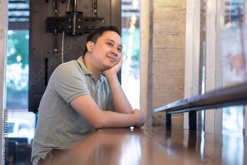 South East Asian man sitting in indoor cafe, contemplating with a smile, hand on chin, eyes looking sideways and upwards as if looking into the distance, wearing green polo shirt.