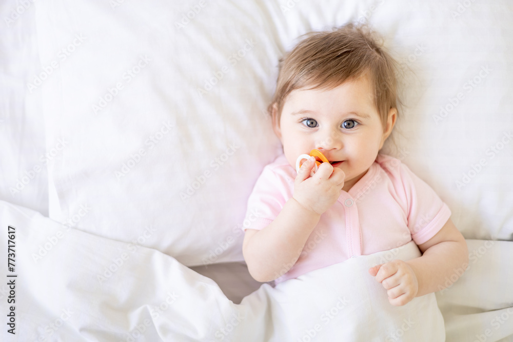 baby newborn girl on a white crib in a bright bedroom close-up in pink clothes sucking a pacifier, place for text, top view