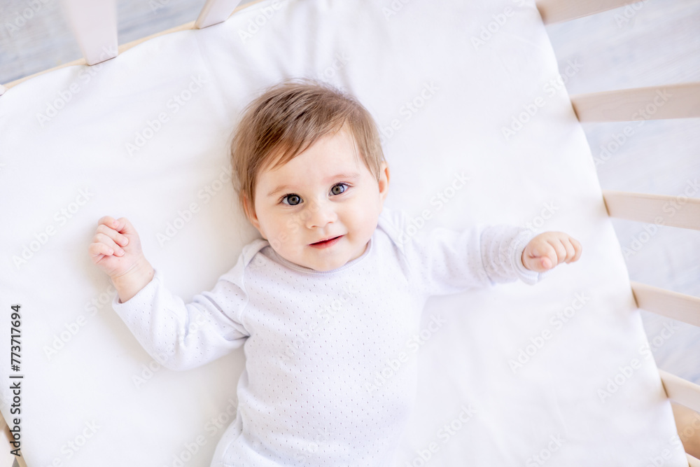 happy baby newborn girl on a white crib in a bright bedroom close-up in white clothes smiling or laughing, place for text, top view
