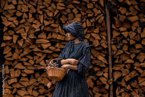 Obraz na plátne A pioneer woman in a dark blue dress and bonnet standing with a basket of flowers against a wood pile