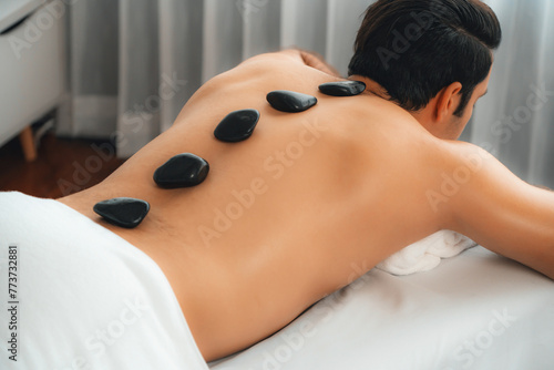 Hot stone massage at spa salon in luxury resort with day light serenity ambient, blissful man customer enjoying spa basalt stone massage glide over body with soothing warmth. Quiescent