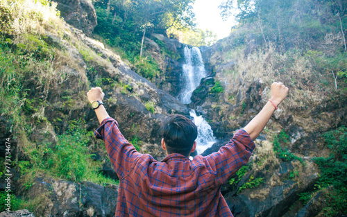 Stylish young traveler or vlogger holding camera and shooting himself, recording video report of his journey for his vlog, standing at Iruppu waterfalls in Coorg or Madikeri, Karnataka, India.  photo