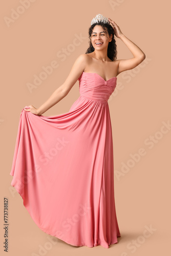Happy young African-American woman in stylish pink prom dress and tiara on brown background