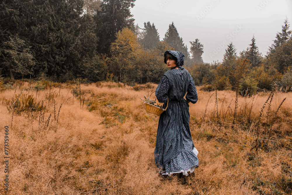 Pioneer woman in a dark blue dress walking in a grassy meadow. Foraging basket. Historical reenactment.