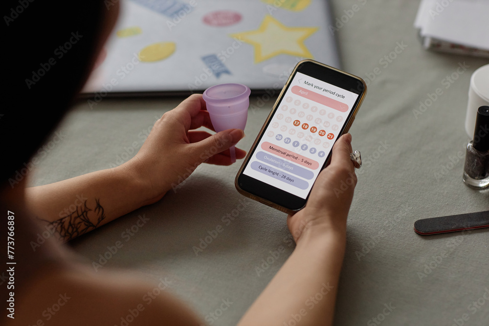 High angle view of unrecognizable young woman holding silicone menstrual cup checking period calendar on smartphone High angle view of unrecognizable young woman holding silicone menstrual cup checking period calendar on smartphone