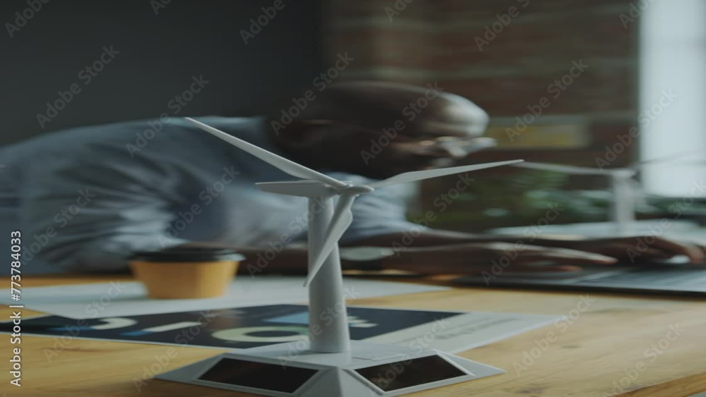 Close-up view of windmill turbine model on office desk, male engineer working on laptop in defocused background. Vertical clip Close-up view of windmill turbine model on office desk, male engineer working on laptop in defocused background. Vertical clip