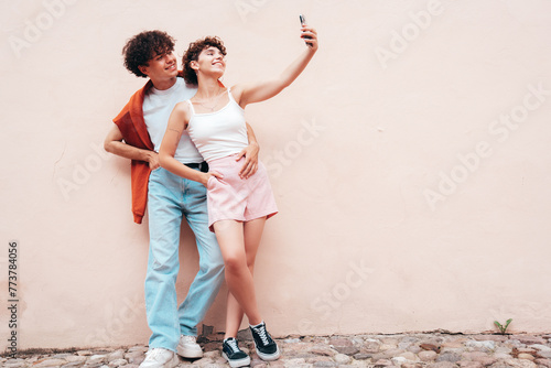 Young smiling beautiful woman and her handsome boyfriend in casual summer clothes. Happy cheerful family. Female having fun. Couple posing in street at sunny day. Near  wall. Take selfie photos photo