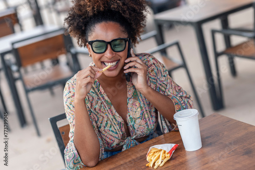 Happy woman talking on smart phone and eating french fries at sidewalk cafe photo