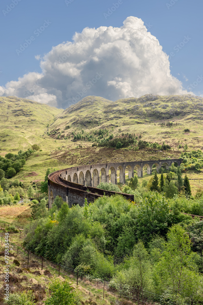 The historic Glenfinnan Viaduct arcs through the verdant Scottish landscape, its ancient stones contrasting the vibrant natural tapestry under a vast, cloud-filled sky