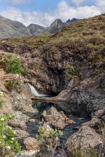 A crisp waterfall spills into the tranquil Fairy Pools of the Isle of Skye, nestled within a rugged landscape with the majestic Cuillin peaks in the backdrop