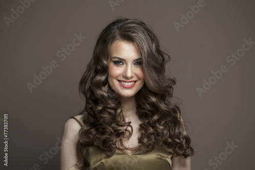 Beautiful woman with long healthy wavy hair, cute smile and make-up looking at camera on brown studio wall background, fashion beauty portrait