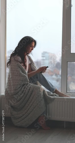 beautiful woman wrapped in a blanket sitting on the windowsill and using phone