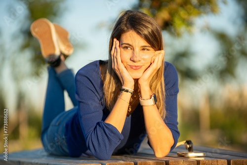 Relaxed woman looking thoughtful while lying on a park bench.