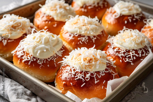 Freshly baked buns topped with cream cheese and shredded coconut on a baking tray with parchment photo