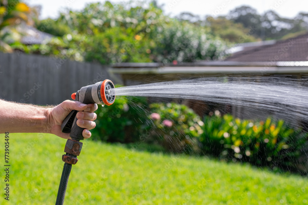 Garden hose with nozzle. Man's hand holding spray gun and watering plants, spraying water on grass in backyard.