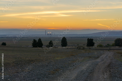 Evening sunset view. An earthen road, trees, a series of mountains, an orange sky. Industrial pipes with smoke and tower. Georgia.