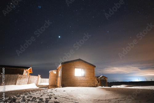 A cozy wooden cabin in Iceland, illuminated against a starry night sky, offers a warm haven amidst the snowy landscape photo
