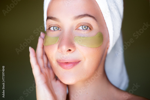 A serene woman with a towel on her head and green hydrogel eye patches smiles softly against a dark backdrop. photo