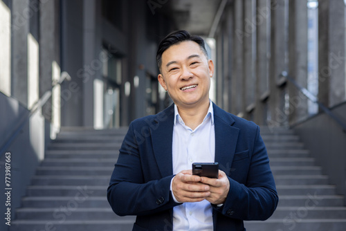 Close-up portrait of a young Asian man in a suit standing outside an office building, holding a phone and smiling at the camera