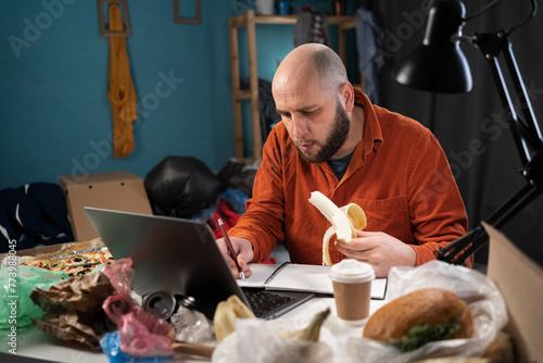 Freelancer working at home in dirty cluttered room, man using laptop computer eating banana at workplace