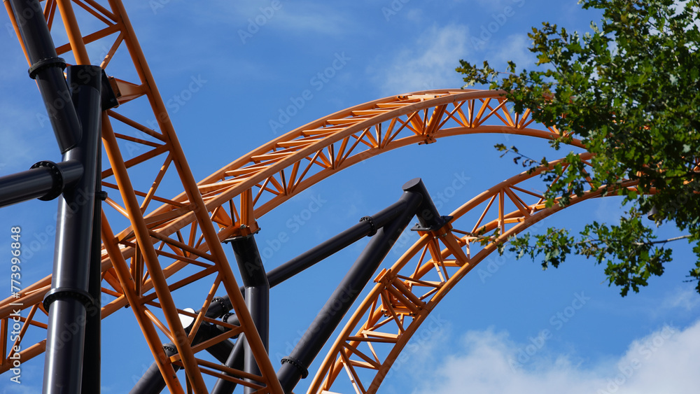 Orange-coloured roller coaster tracks of an extreme roller coaster, crossing under a blue sky and a tree in the foreground Orange-coloured roller coaster tracks of an extreme roller coaster, crossing under a blue sky and a tree in the foreground