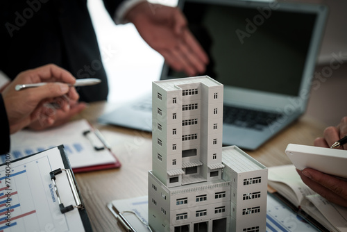 A professional businessman in a suit conducts a meeting at a desk while showcasing a model of a condominium or apartment building, discussing real estate development plans.
