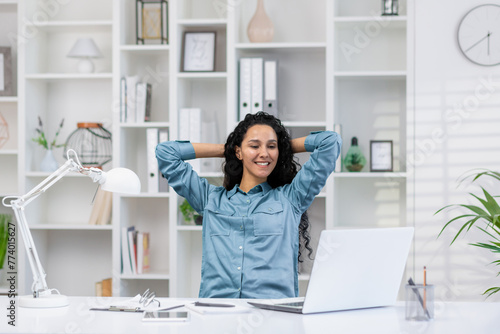 A relaxed Hispanic woman takes a break while working remotely in her bright home office, embodying work-life balance.