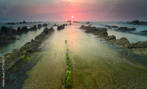 A fiery sunset casts a warm glow over the moss-covered rocks and silken waters of Playa de Barrika in Bilbao, creating a surreal seascape photo