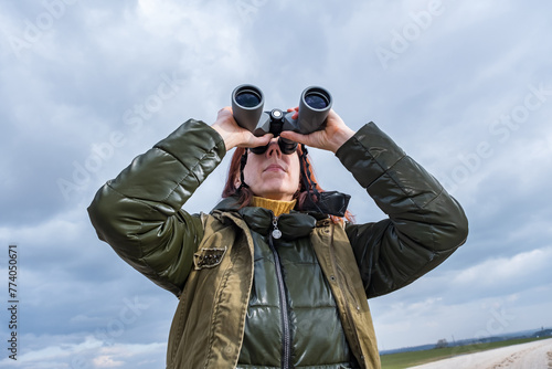 female ornithologist birdman watches birds with binoculars on dirt gravel road against a background of a stormy sky photo