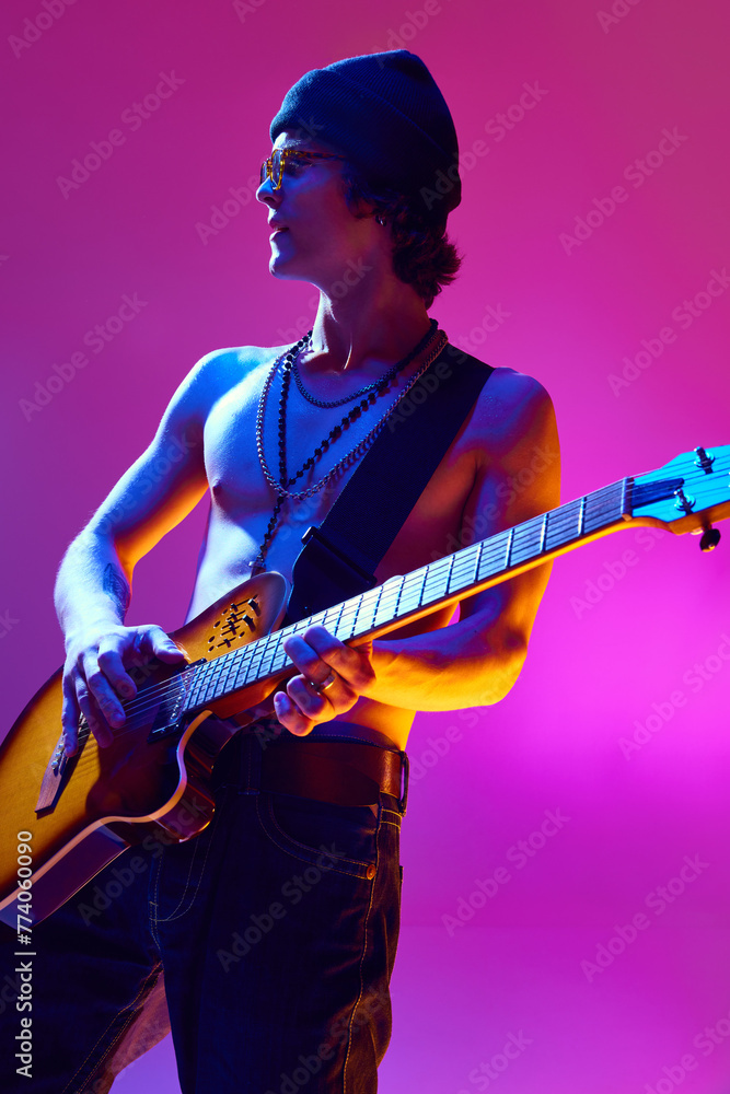 Obraz premium Handsome shirtless young man in sunglasses and hat playing electric guitar against pink background in neon light. Concept of music, talent show, performance, concert, festival, instruments