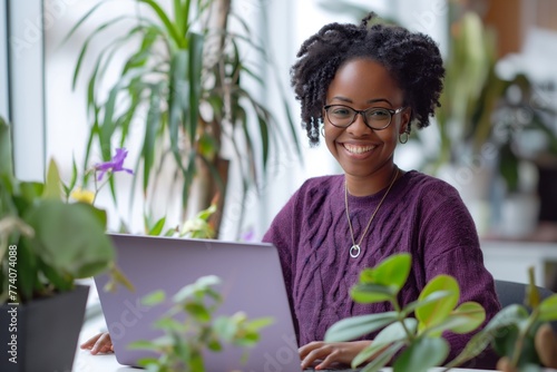 An attractive black woman of 30 years old, wearing a purple sweater and glasses, smiling and looking happy, sitting at a table with a laptop surrounded by plants, and working in a home office © Vadim