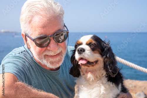 Portrait of old attractive senior bearded man sitting close to the sea with his cavalier king charles dog. Best friend forever concept. Horizon over water
