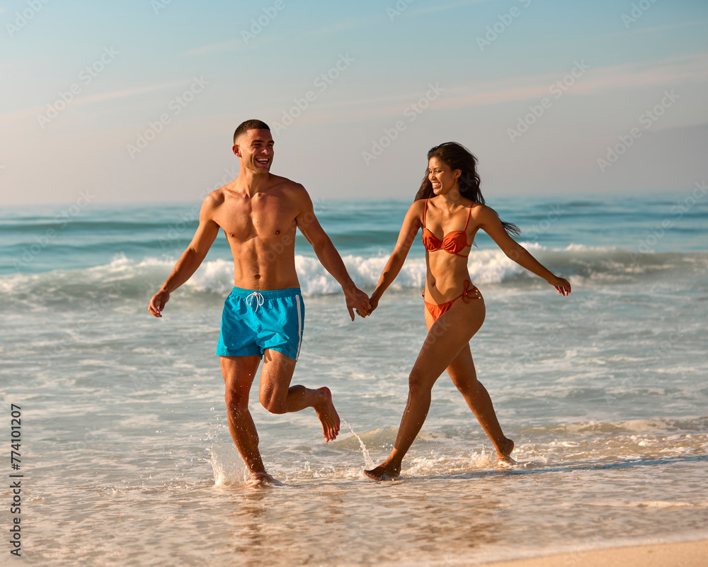 Couple In Swimwear On Vacation Holding Hands Running Along Beach Shoreline 
