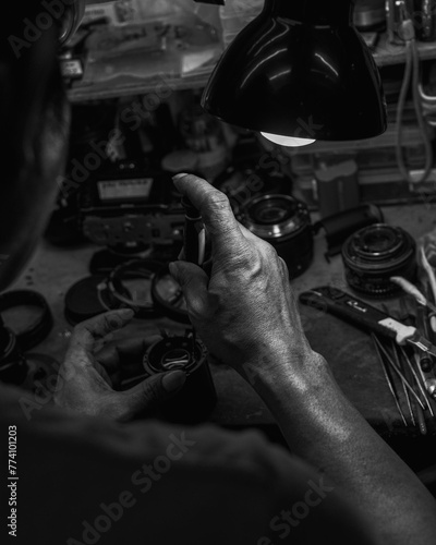 Vertical grayscale of human hands repairing camera parts under a table lamp light