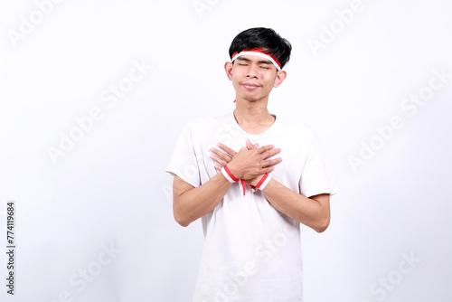 Relieved Young asian boy with red white ribbon celebrating Indonesia independence day while holding his chest and closing eyes photo