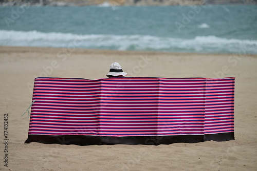 A man with a sunhat behind a big, red textile wind blocker at the beach of Benidorm-Spain on a stormy April day.