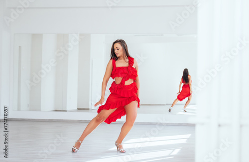 Graceful woman dressed in red Latin dancing dress doing elegant dance movement in white color big hall with big mirror wall. People's expressions during dancing, beauty of woman's body concept image