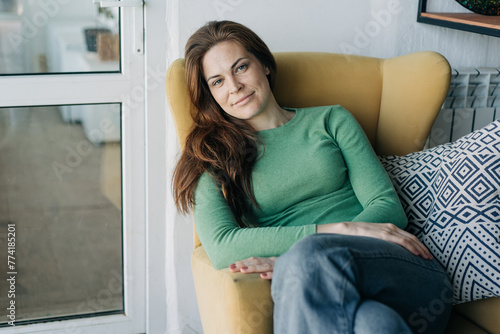Fotografie A gorgeous long-haired red-haired woman sitting in a chair calmly looks at the camera