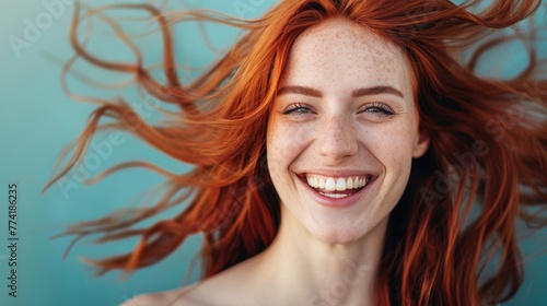 A young woman with flowing red hair and freckles smiles brightly against a blue background, her joy contagious. photo