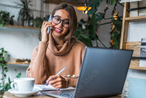 Freelancer woman sitting in a coffee shop with a laptop on the table while making notes and talking on cellphone. Focused businesswoman in casual outfit drink coffee while working remotely in cafe
