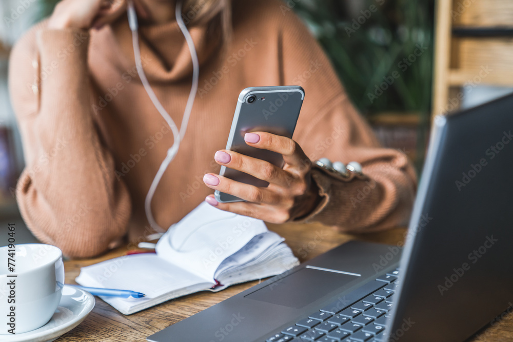 Close up shot of smartphone being used by a woman in earphones sitting at table. Woman in casual outfit working remote on laptop, make notes, listening business podcast while have coffee break on cafe