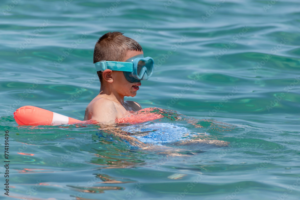 Boy in swim goggles resting after swimming lesson, using floating equipment for safety. Child enjoying water sports while relaxing on summer beach vacation