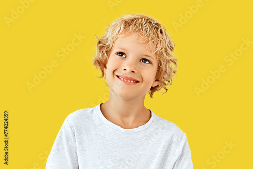 A cheerful young boy with curly blonde hair looks up thoughtfully, wearing a white shirt against a bright yellow background.