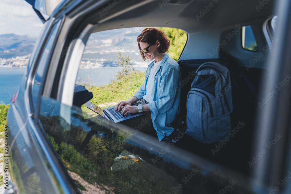 Young woman sitting in the open trunk of a car overlooking the sea and using a laptop, freelancer, remote work and summer vacation auto travel