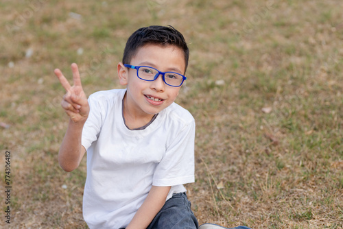 Hispanic boy playing in the field - 8 year old boy sitting on the grass in the park - Latin boy playing and having fun in nature - playing and learning - boy with white t-shirt